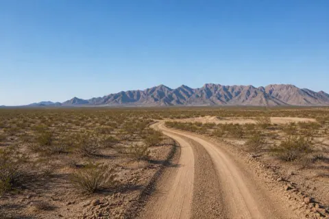 Fresh sandy access drive curving through creosote flats toward a distant homesite pad, the Gila Mountains ahead