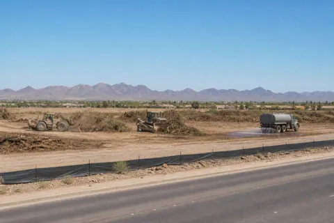 Unbranded machines opening commercial acreage along a Yuma arterial, mulched mesquite in windrows while a water truck wets the pad