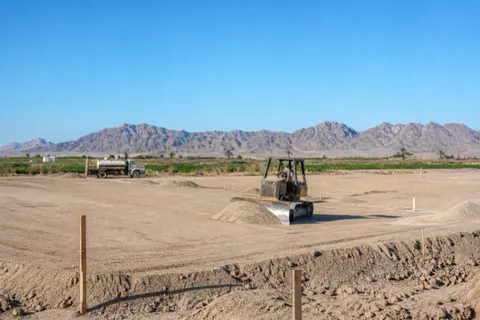 Compact dozer feathering pale sand to level on a Yuma homesite pad, string lines and grade stakes already set