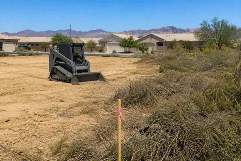 Half-cleared residential lot in a growing Yuma subdivision, smooth pale sand meeting untouched native scrub, pink flagging on the corner stakes